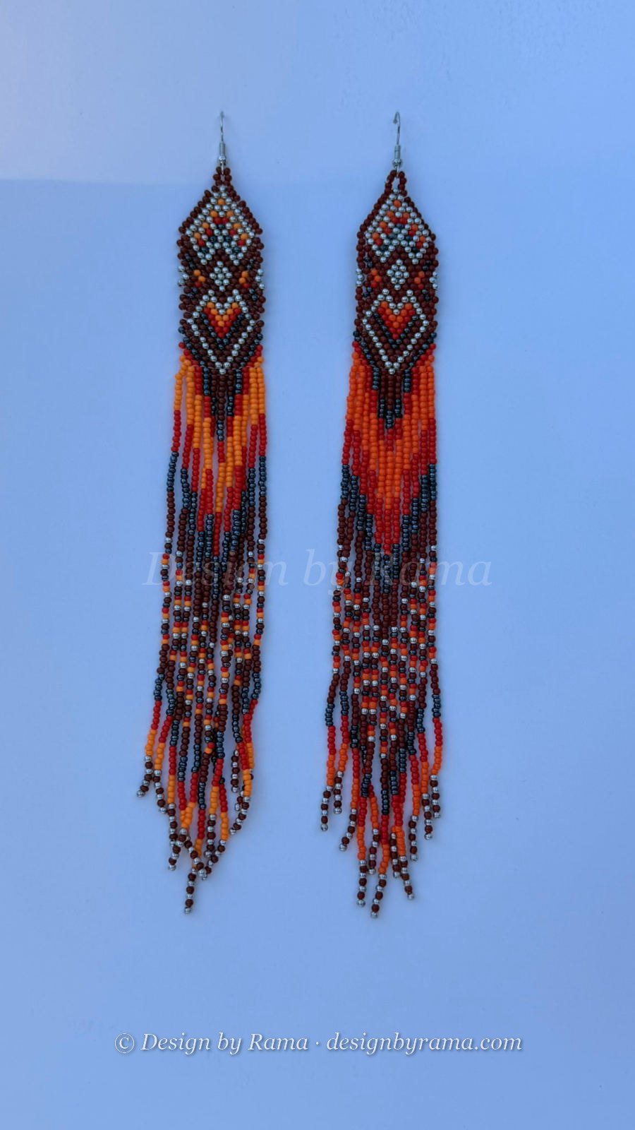 Very long fringe drop earrings with geometric diamond chevron header panel in oxblood, burnt orange, tangerine, pale silver-grey, and white seed beads, extra-long fringe repeating the same chevron color pattern with silver metallic accent beads at the tips, silver fish-hook closures.